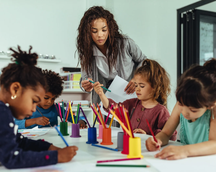 Teacher helping a young child start drawing by giving them a pencil, while other children work at the classroom table