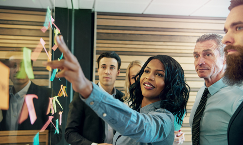 Young female apprentice leading a meeting and presenting her ideas to a diverse team in front of a wall of sticky notes, highlighting collaboration, apprenticeships, and workforce upskilling.